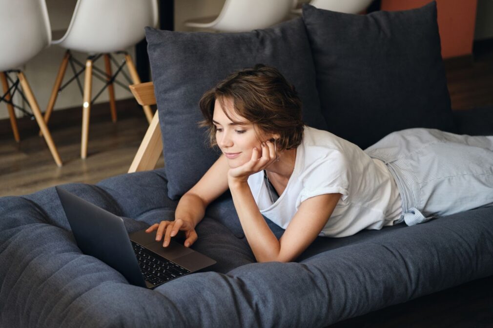 A woman is lying on the couch playing on the computer